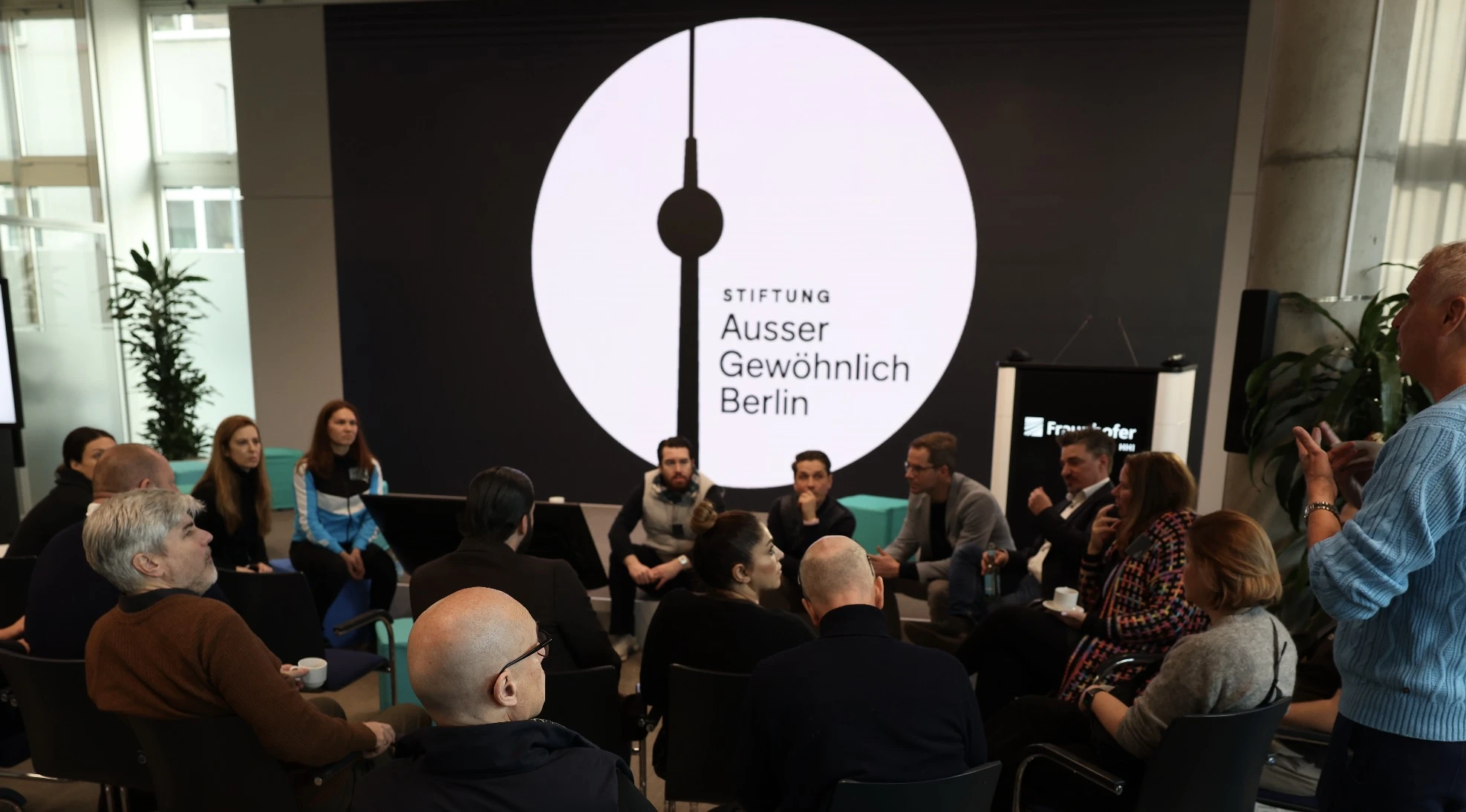 Menschen im Gesprächskreis bei einem Salon, vor dem großen Logo der Stiftung AusserGewöhnlich Berlin